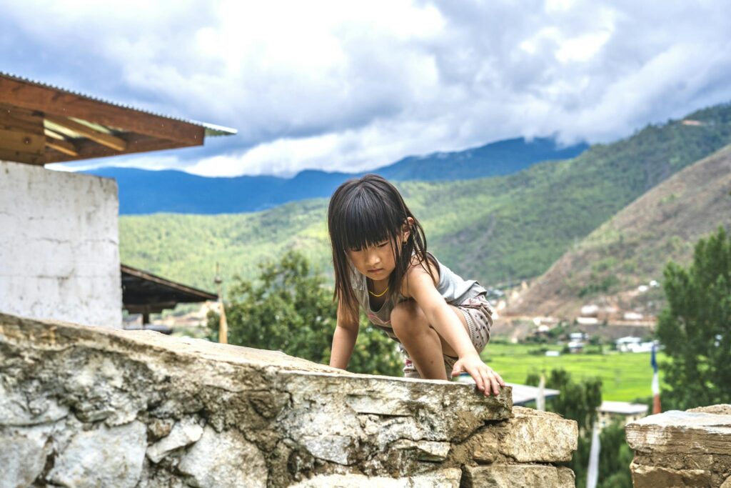 A young child climbing a stone wall in a scenic mountain village setting.